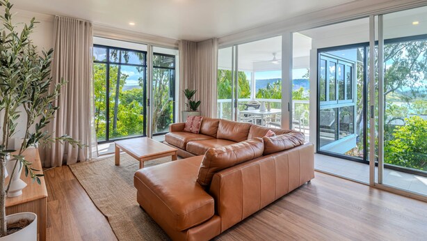 A modern living room with a brown leather sofa, a wooden coffee table, and a large window offering a view of greenery and a balcony. - Oasis 2, Beautiful Apartment on Hamilton Island (Whitsundays)