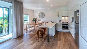 A modern kitchen with a central island, wooden flooring, and a dining area with white chairs. - Oasis 2, Beautiful Apartment on Hamilton Island (Whitsundays)