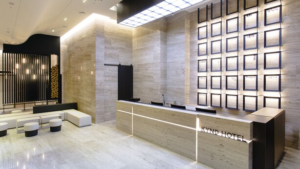 Un espace d’accueil moderne, avec un comptoir en bois, un coin salon et un mur où sont affichés les noms de différentes villes.