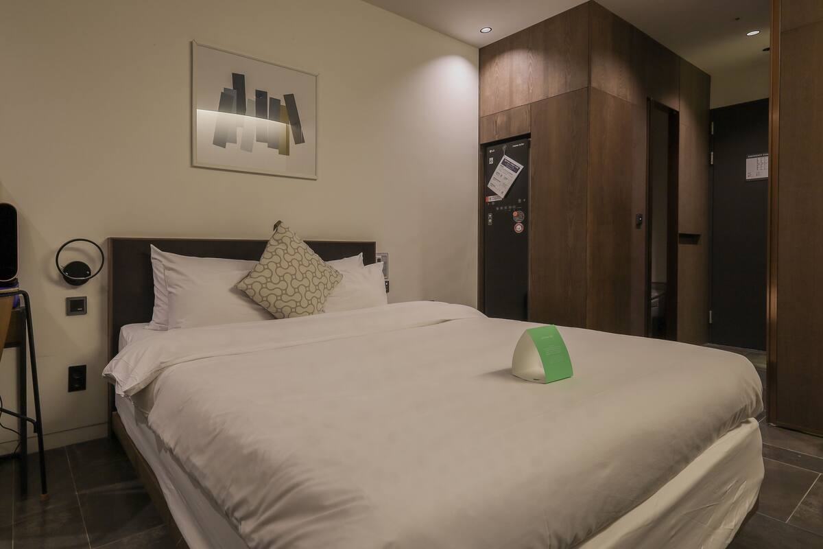 A hotel room with a bed, a desk, a chair, a bedside table, a mirror, a wall-mounted control panel, and a door.
