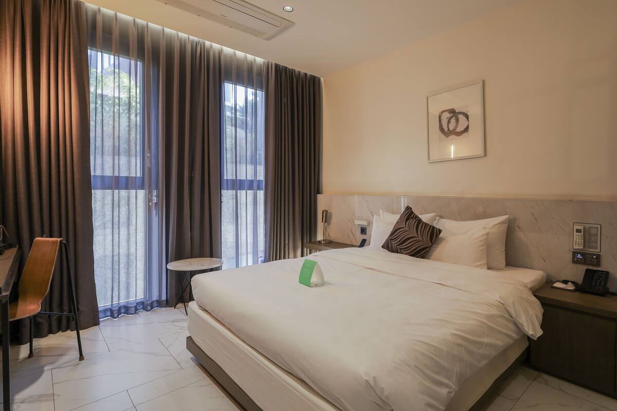 A hotel room with a large bed, a desk with a chair, a small table, and a window with curtains.