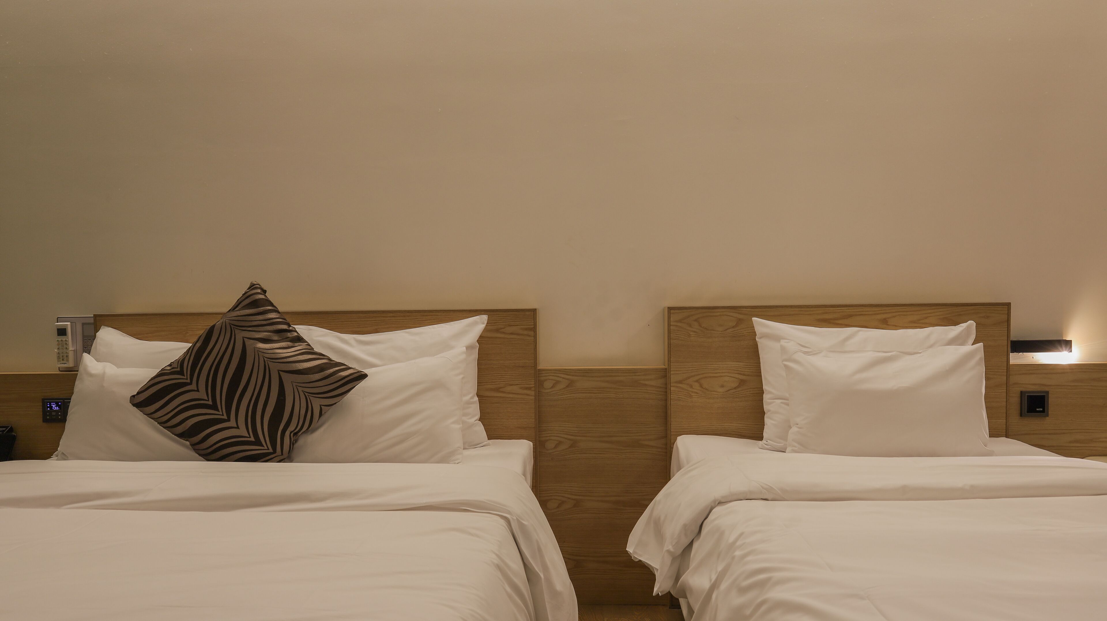 A hotel room with two beds, each with white bedding and a wooden headboard. There is a telephone and a control panel on the wall.
