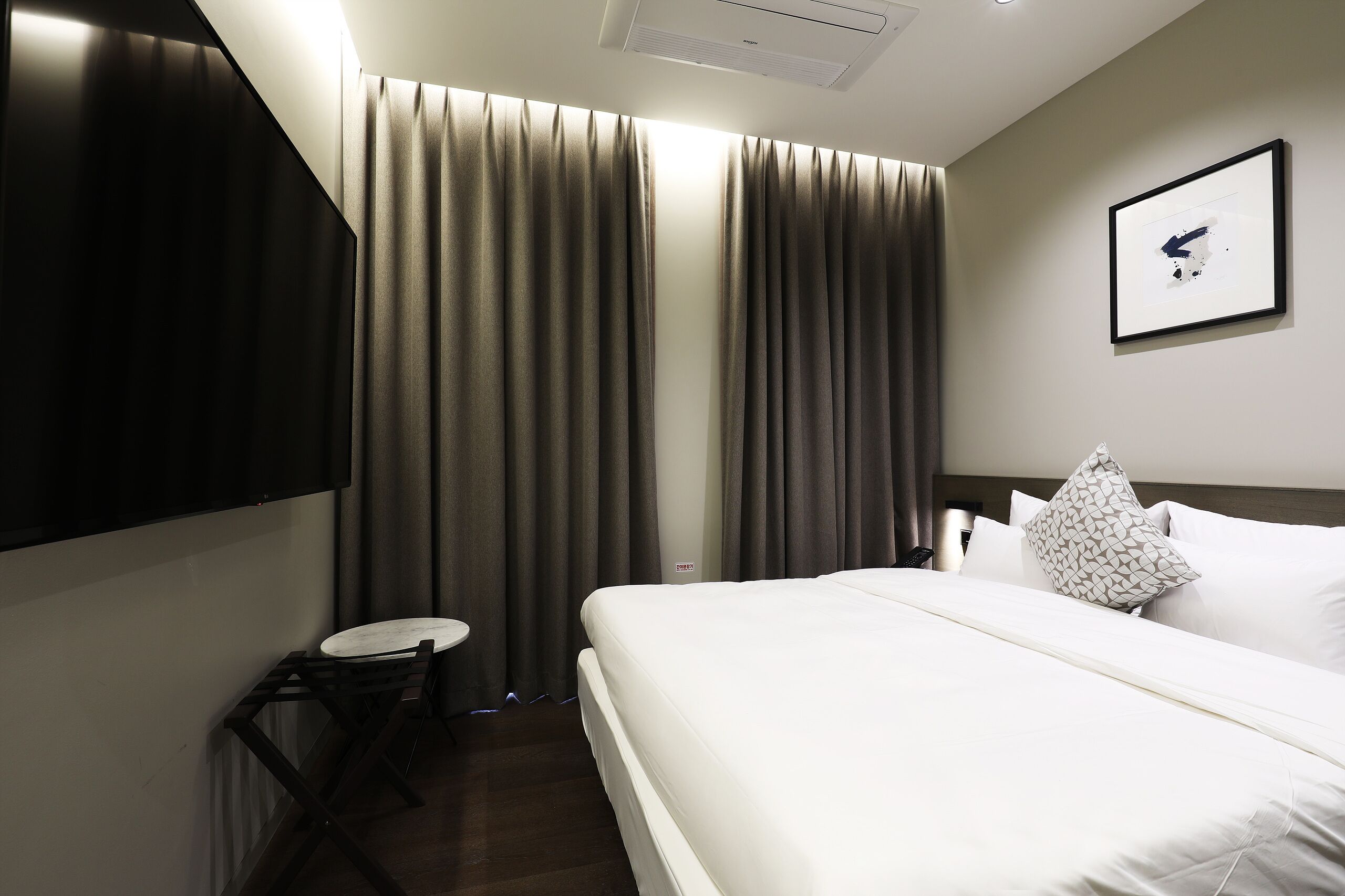 A hotel room with a bed, a flat-screen TV mounted on the wall, a small round table with a chair, and a large window with curtains.
