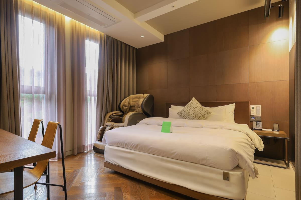 A hotel room with a bed, a massage chair, a table, and a bedside table with a phone and a digital clock.