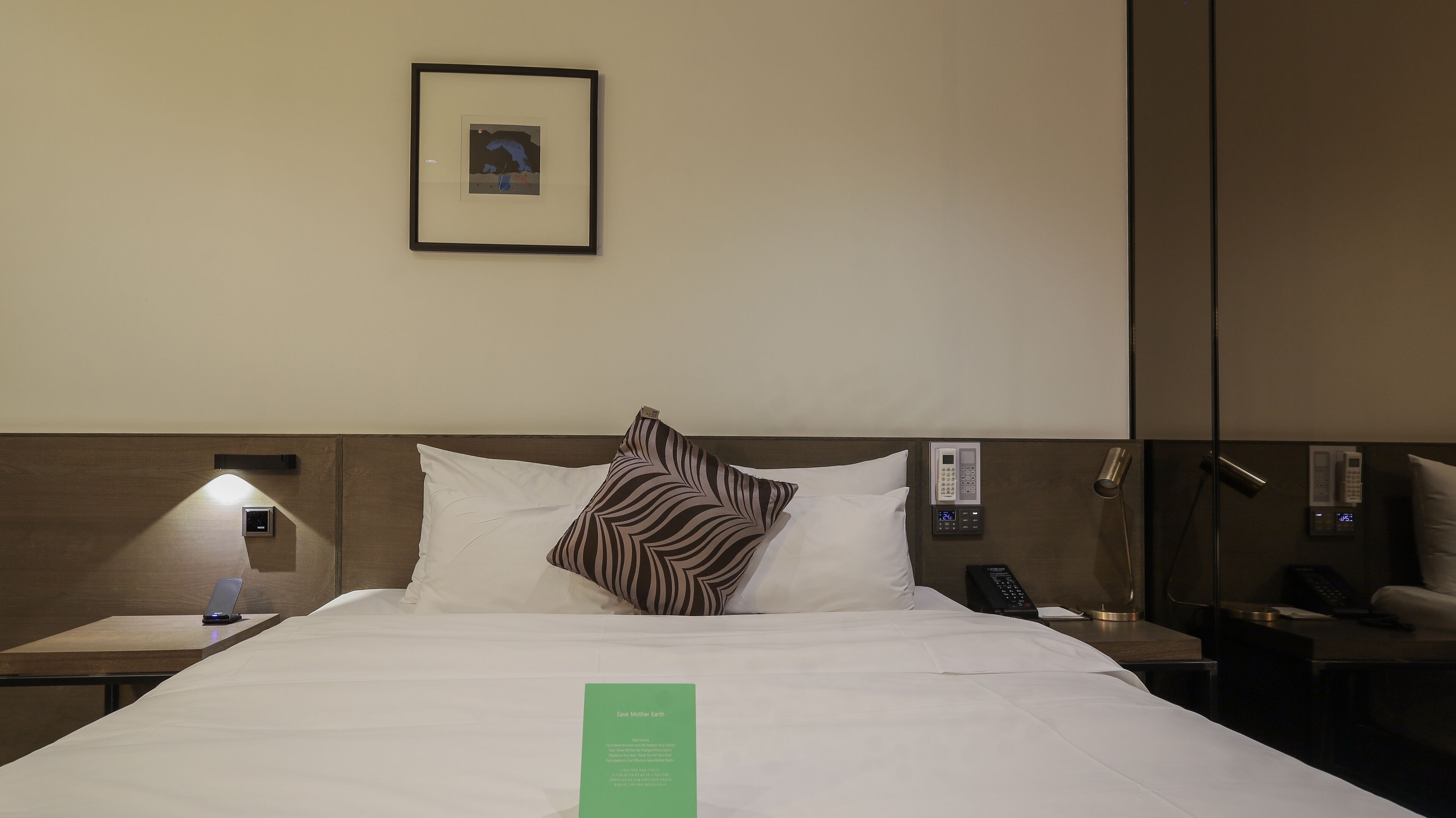 A hotel room with a neatly made bed, a framed picture on the wall, a bedside table with a lamp, and a bedside phone.