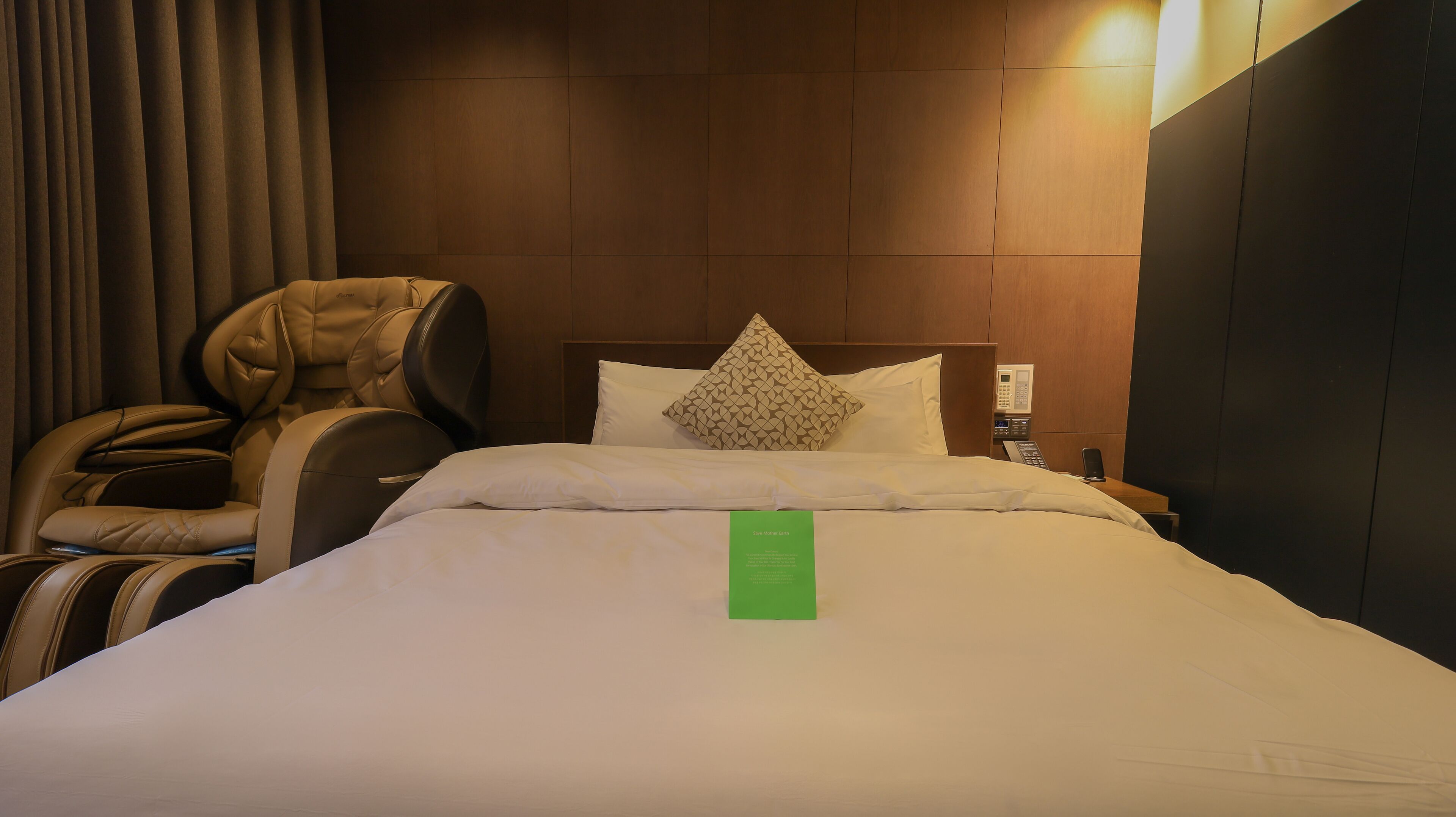Une chambre d’hôtel avec un lit, un fauteuil massant, un oreiller, une table de chevet avec un téléphone et un panneau vert.