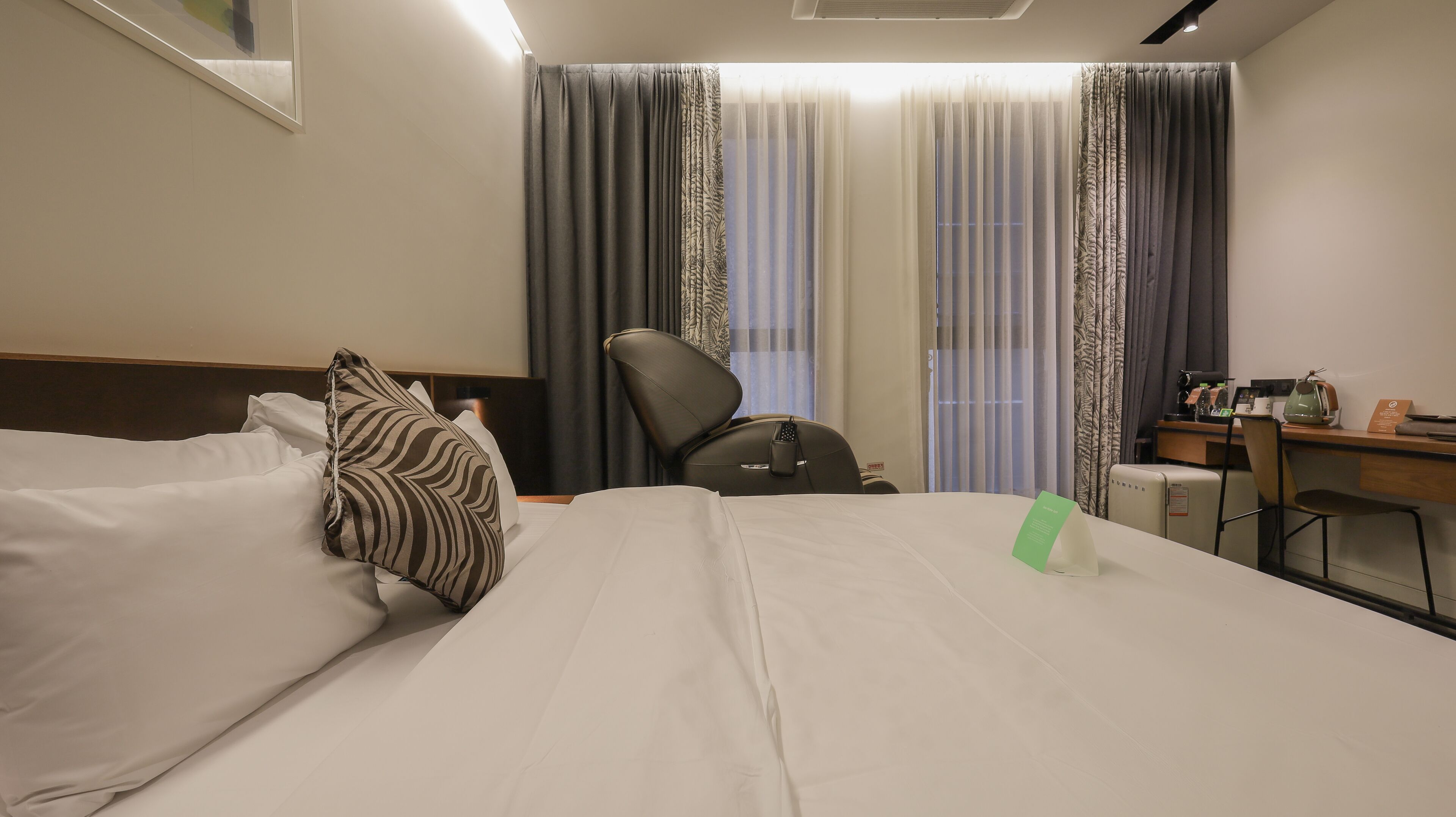 A hotel room with a bed, pillows, a desk, a chair, and a window with curtains.