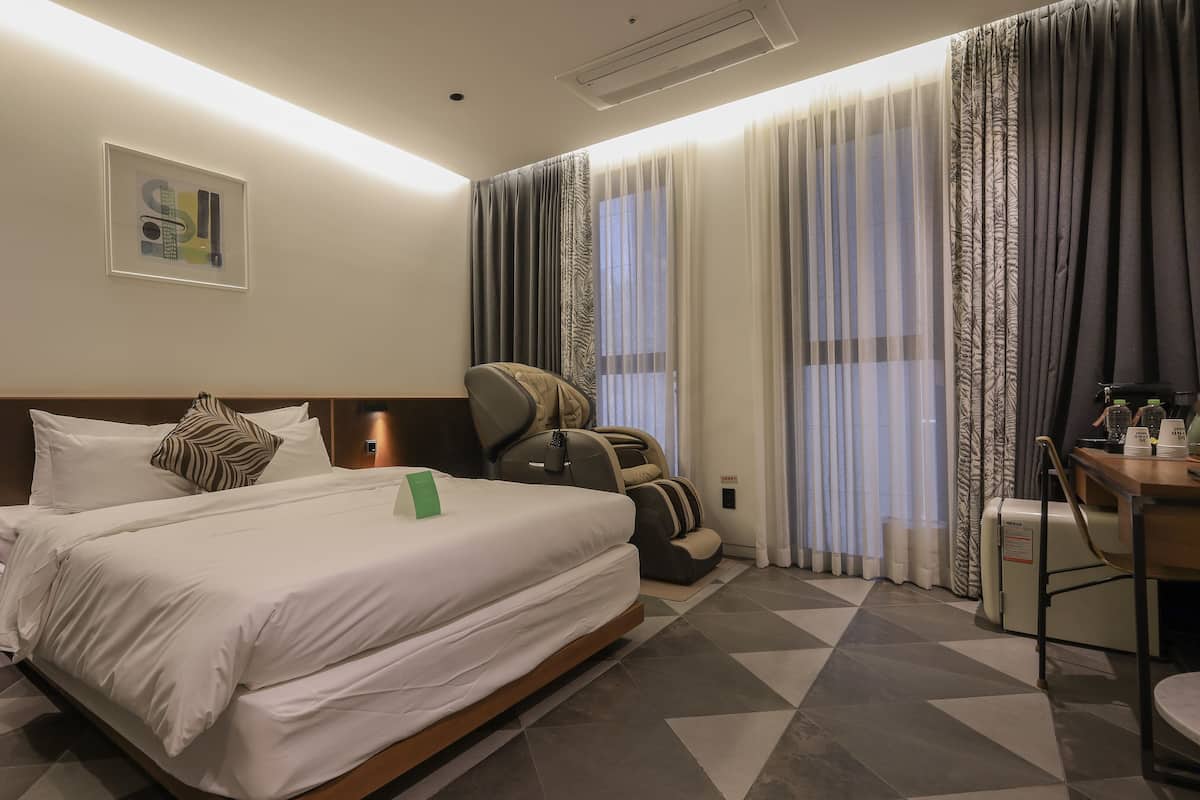 A hotel room with a bed, a massage chair, a desk with a coffee machine, and a window with curtains.