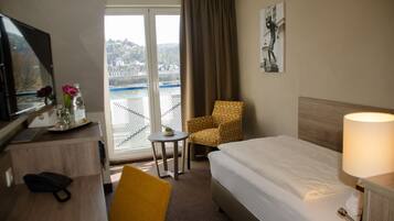 Una habitación de hotel con una cama, un escritorio con una computadora, una silla, una mesita con una planta y vistas a un cuerpo de agua a través de una ventana.