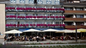 Un hotel con varios balcones decorados con flores rosadas, mobiliario exterior con sombrillas, y un cartel que indica un restaurante en la planta baja.