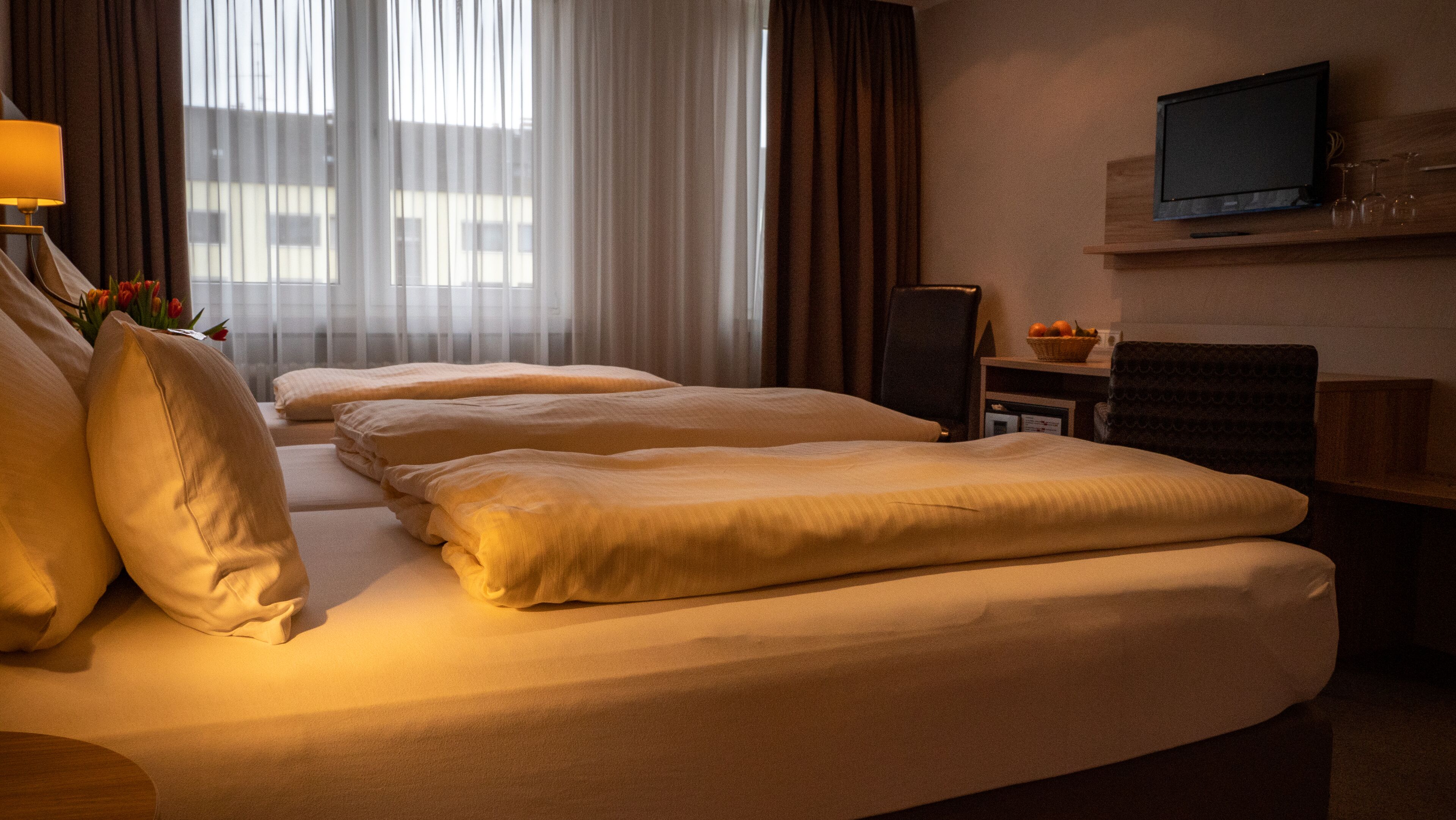 Une chambre d’hôtel avec deux lits, un téléviseur fixé au mur et un bureau sur lequel se trouve un panier de fruits.