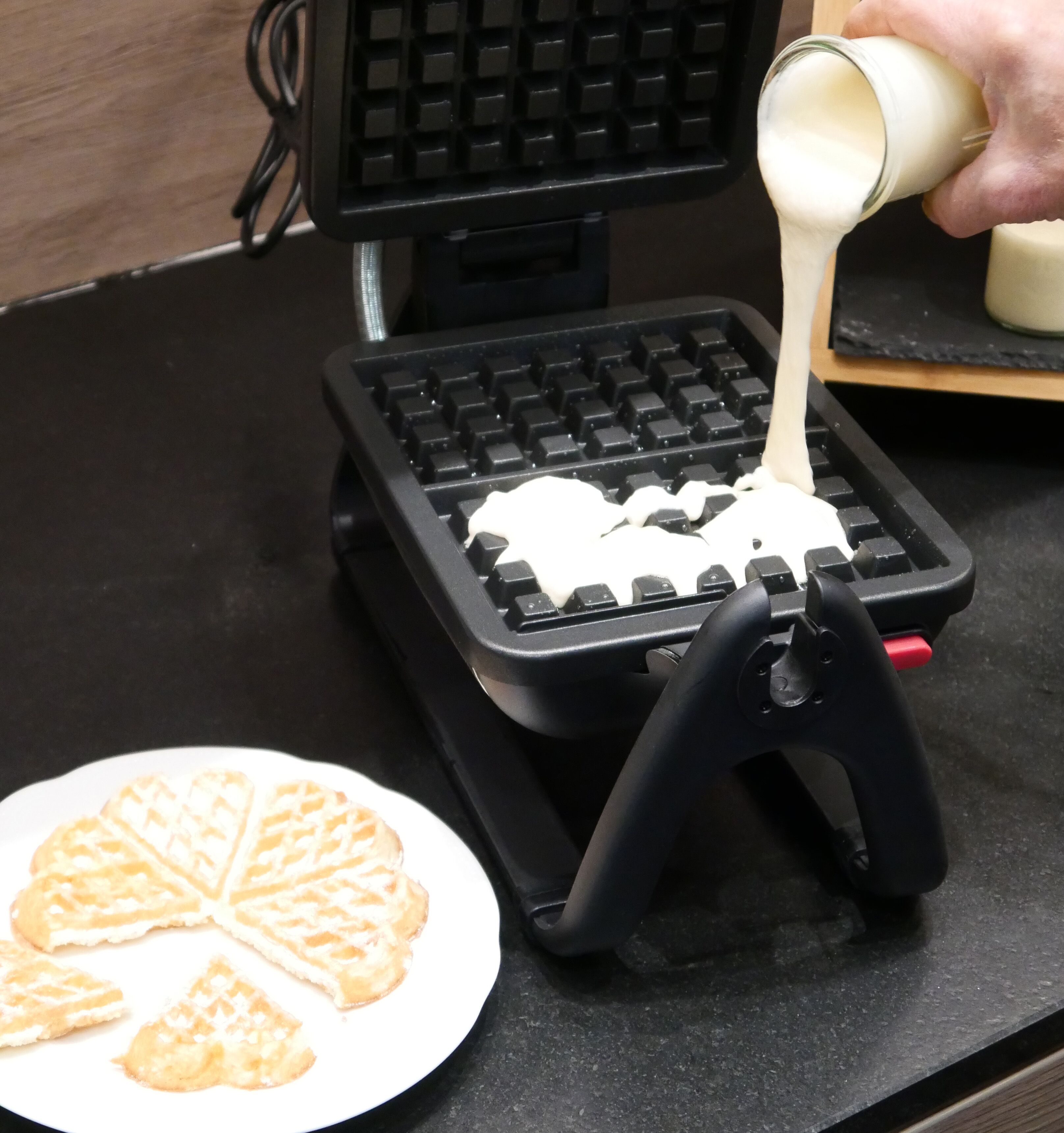 A waffle iron on a black countertop with a hand pouring batter onto it.