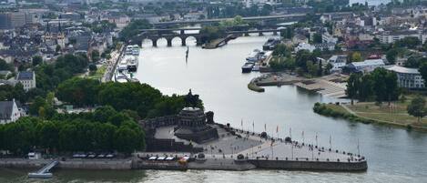 Vista de la ciudad a la orilla del río, con un puente histórico, un paseo marítimo y una gran plaza abierta con banderas.