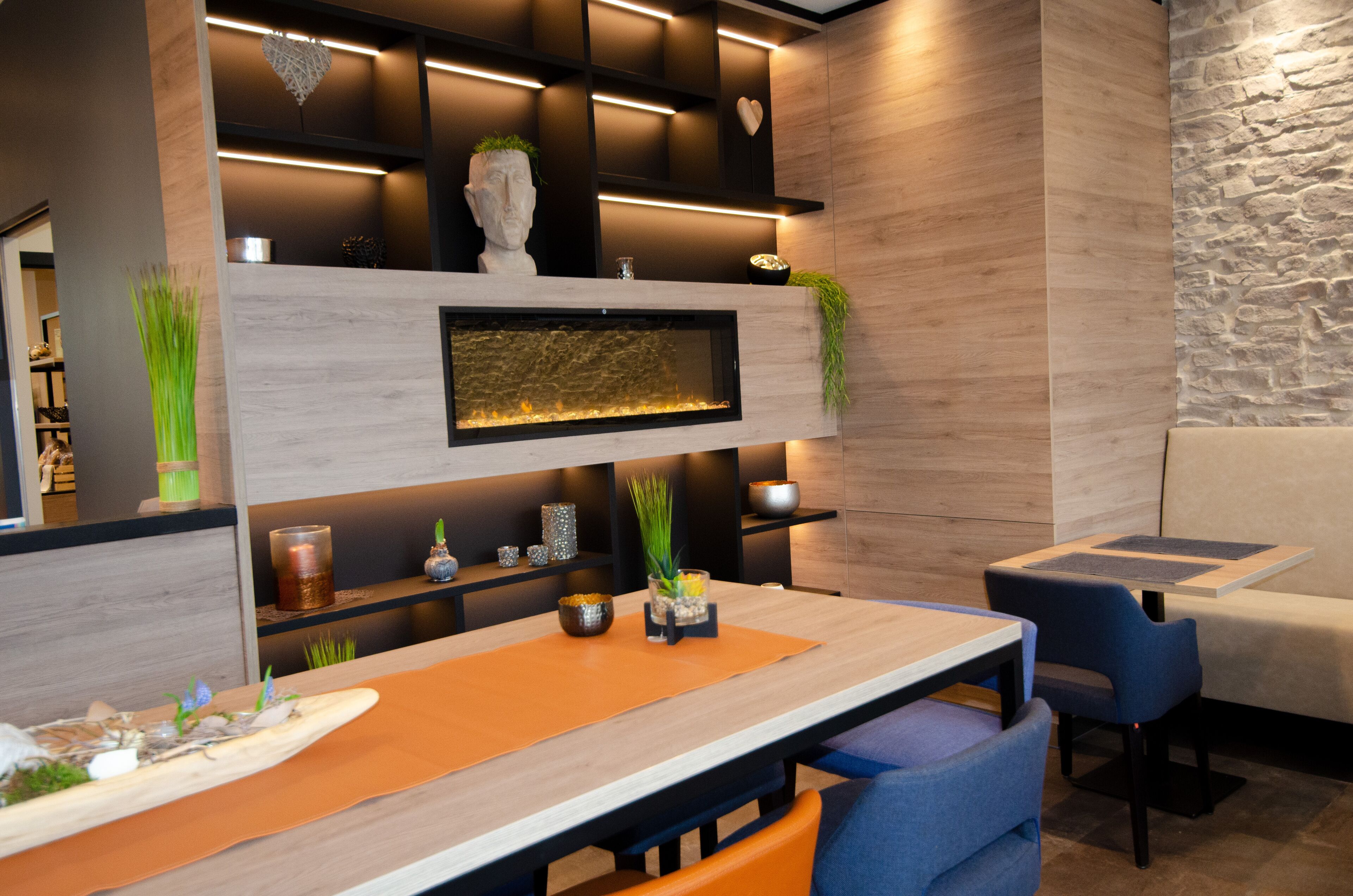 A modern interior with a dining area featuring a wooden table, orange placemat, and blue chairs. There is a built-in shelf with decorative items and a small waterfall feature.