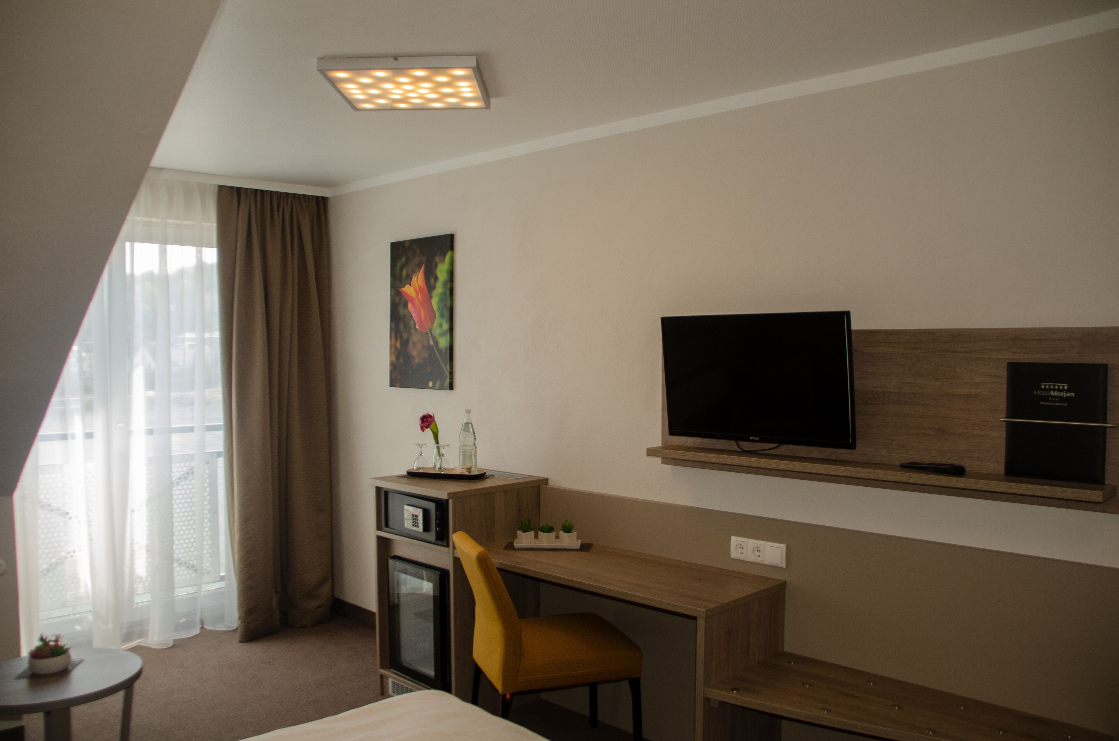 Habitación de hotel con cama, escritorio con silla, televisor, refrigerador pequeño y ventana con cortinas.