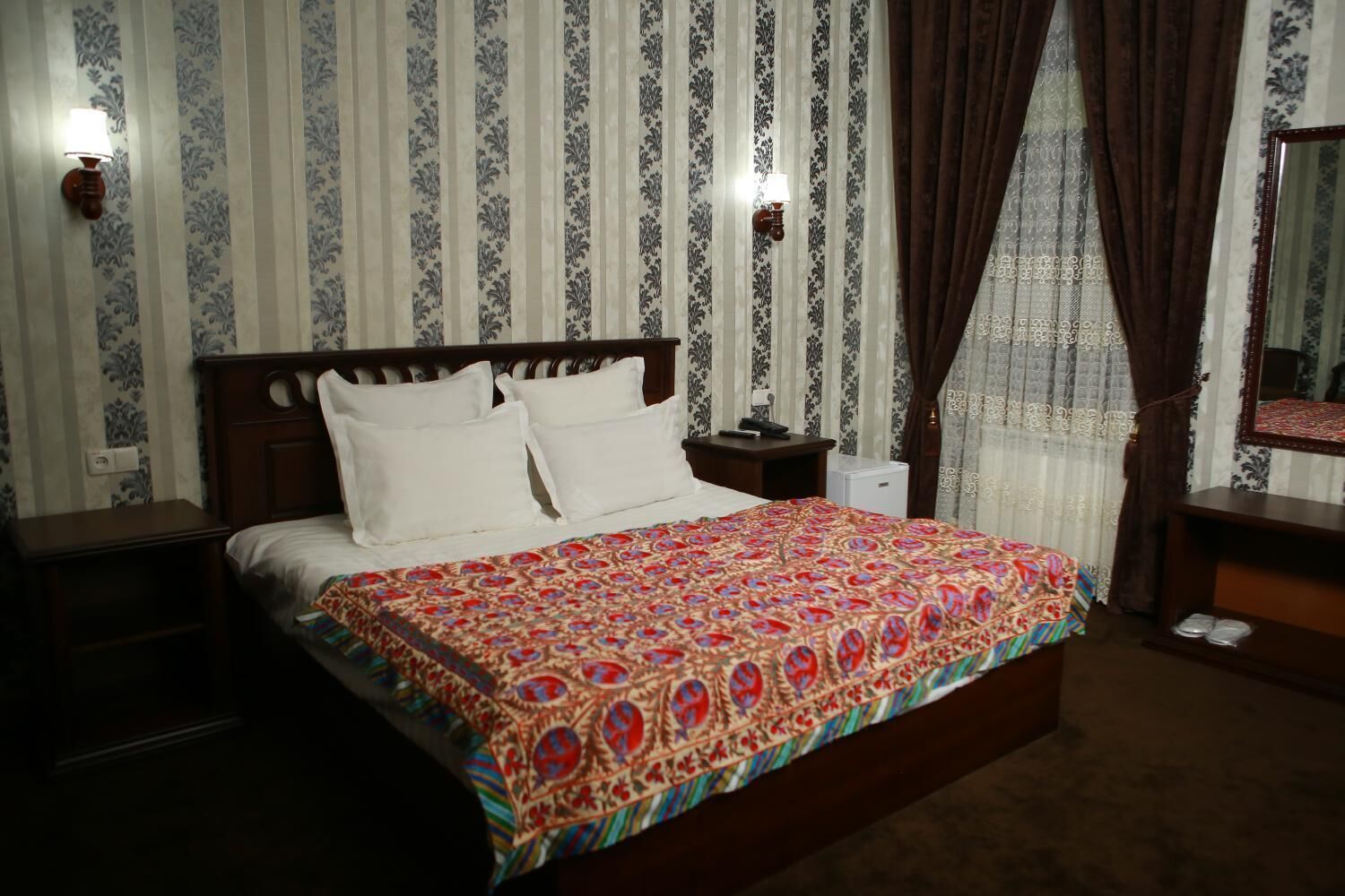 A neatly made bed with white linens and a colorful patterned bedspread. There is a wooden nightstand with a lamp, a small bedside table, and a mirror on the wall. The room has striped wallpaper and a window with brown curtains.