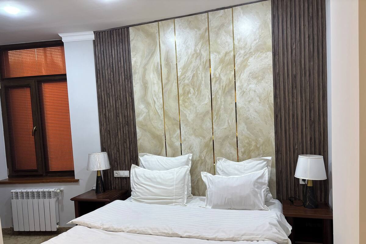 A neatly made bed with white linens, two bedside tables with lamps, a radiator, and a window with blinds.