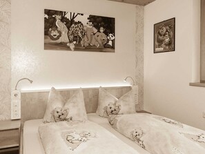 Une chambre à coucher avec un lit agrémenté d’une couette et de coussins à motifs d’ours, une table de chevet avec une lampe et un mur orné de deux tableaux encadrés.