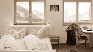 A bedroom with a bed, a desk, and a chair. There are windows with a view of mountains. - Apartment 2 - Apartments Madeleine (Schoppernau)