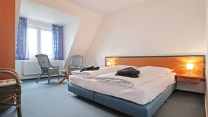 Une chambre d’hôtel moderne avec un grand lit, deux fauteuils, une petite table et une fenêtre avec des rideaux.