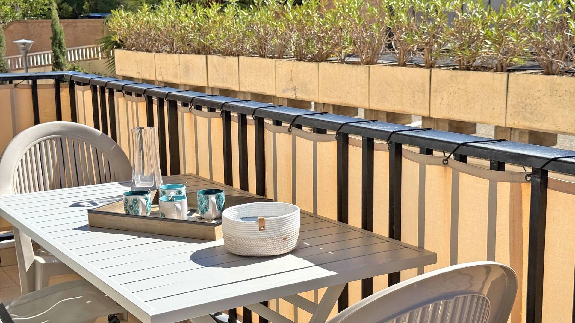 Eine Terrasse mit einem weißen Tisch und Stühlen, einer Ablage mit Tassen und einem Glas sowie einem Pflanzgefäß mit kleinen Sträuchern.