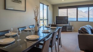 A dining area with a long table set for four, a flat-screen TV, and a view of mountains and water. - Lake and Mountain Ridge - Queenstown Apartment (Queenstown)