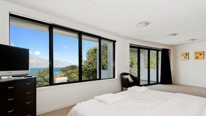 A modern bedroom with a large bed, a TV on a dresser, and a view of a lake and mountains through large windows. - Lake and Mountain Ridge - Queenstown Apartment (Queenstown)