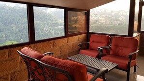 A balcony with a table and two chairs, offering a view of a forested area and a cityscape. - Pinewood Hotel (Jeita)