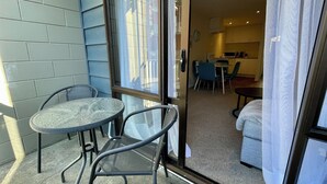 A small balcony with a glass table and chairs, a sliding glass door leading to a compact living area with a kitchenette, a dining table, and a sofa.