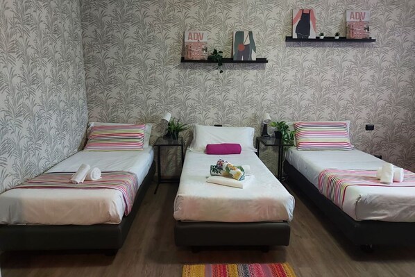 Habitación de hotel con tres camas individuales, cada una con una almohada y una toalla enrollada. Pequeñas mesitas de noche con lámparas y una planta en maceta sobre cada cama. La habitación tiene papel tapiz estampado y un estante con adornos sobre las camas.