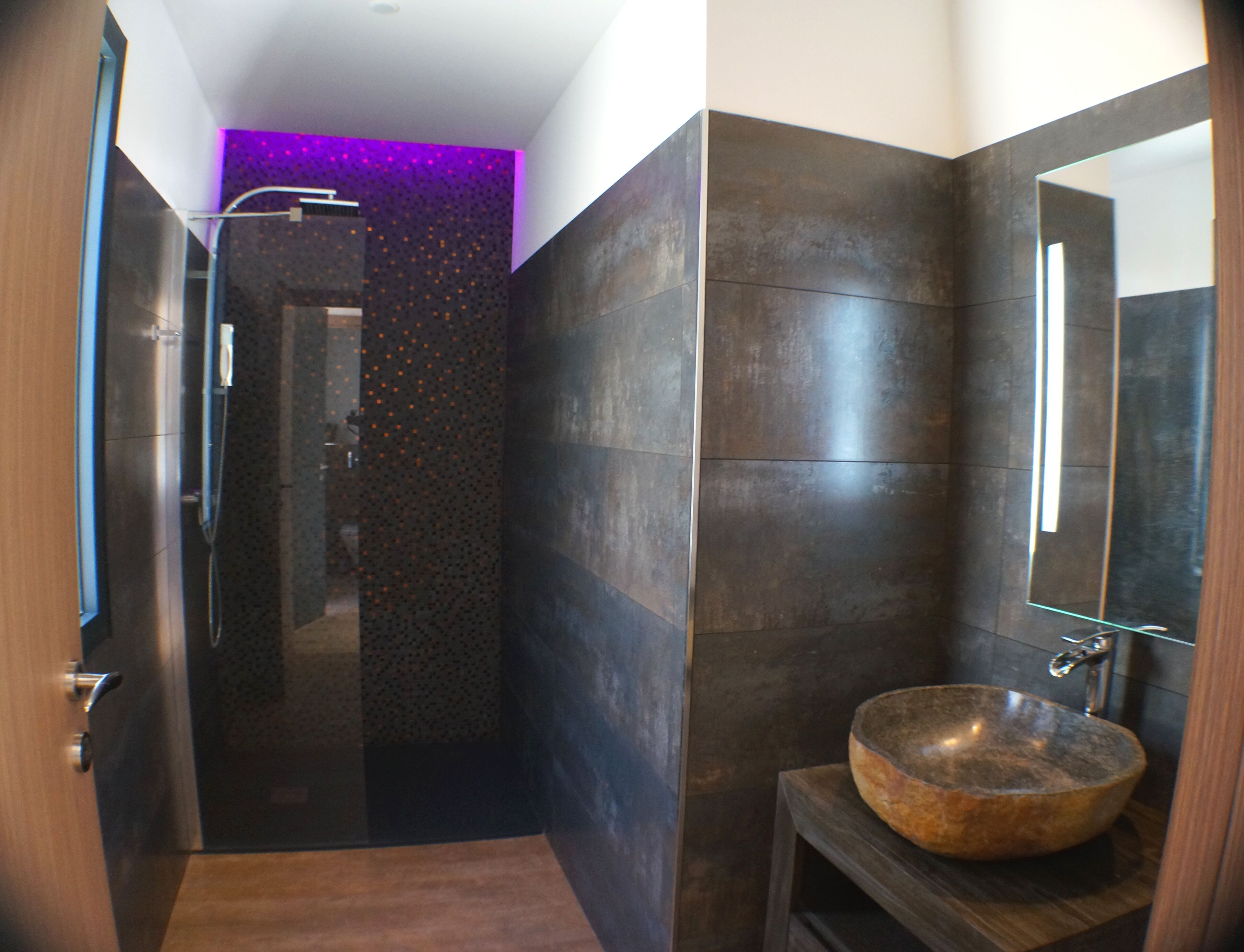 A modern bathroom with dark tiles, a wooden vanity, and a large mirror.