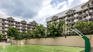Un immeuble à plusieurs étages doté de balcons, d’un terrain de basketball et entouré de verdure.