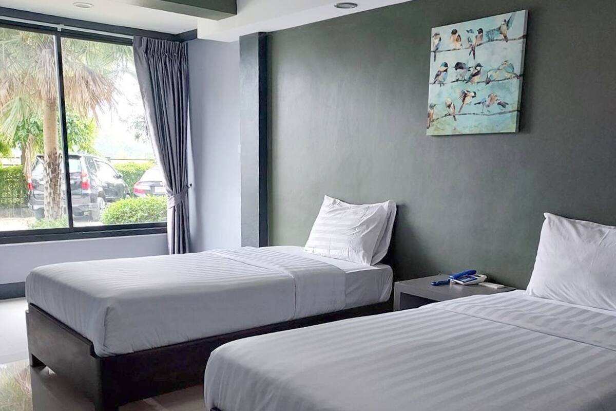 A hotel room with two beds, a window with a view of parked cars and greenery, and a painting of birds on the wall.