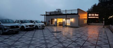 A parking area with several cars, a building with a sign reading "Ell Bee Mussoorie Hills", and a tiled floor.