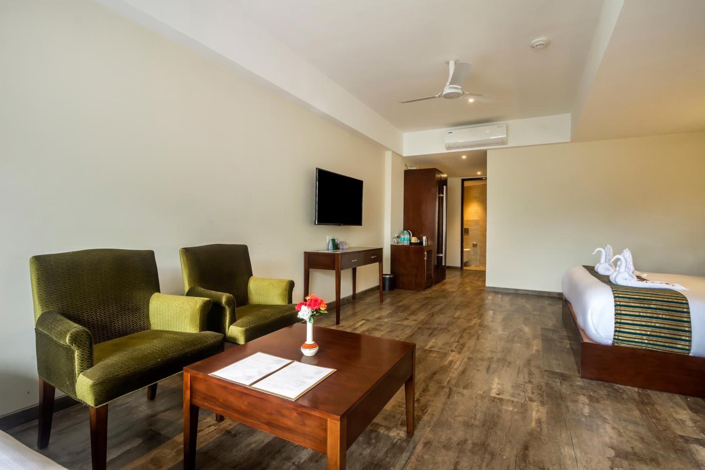 A hotel room with a wooden floor, a bed with white bedding and a green-striped pillow, two green armchairs, a wooden coffee table with papers, a flat-screen TV on the wall, and a small table with a vase of flowers.
