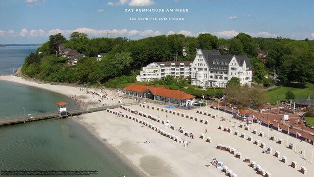 A coastal hotel with a large building surrounded by lush greenery, a beach with sun loungers, and a pier extending into the water. - Large penthouse by the sea - box seat on the fjord - (Glücksburg (Ostsee))