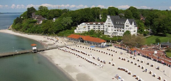 A coastal hotel with a large building surrounded by lush greenery, a beach with sun loungers, and a pier extending into the water.