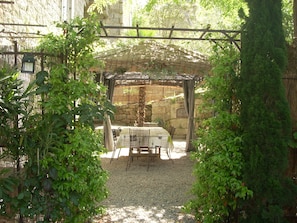 A covered outdoor seating area with a table and chairs, surrounded by greenery and a stone wall.