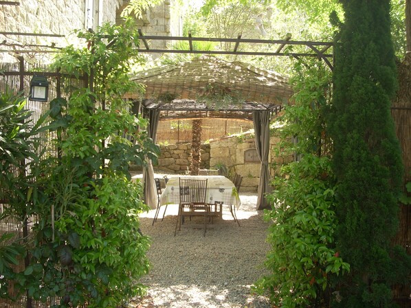 Un espace extérieur abrité, aménagé avec une table et des chaises, entouré de verdure et d’un mur en pierre.