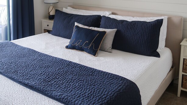 Un lit bien fait, recouvert d’une couette blanche et bleu marine, agrémenté d’un miroir décoratif et d’une lampe de chevet.
