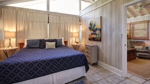 A bedroom with a large bed, two bedside tables, a nightstand, a painting, and a view of a living area through an open door. - Cute, Cozy Beach Cottage/Duplex - One block to the beach! Location is Key! (Isle of Palms)