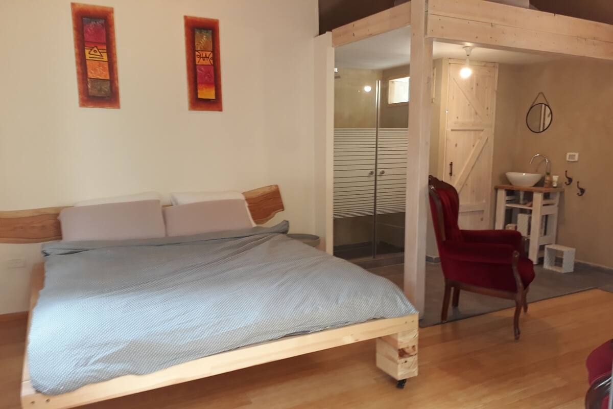 Un dormitorio con cama de madera, una silla roja, una mesita con lavamanos y una puerta corredera que da al baño.