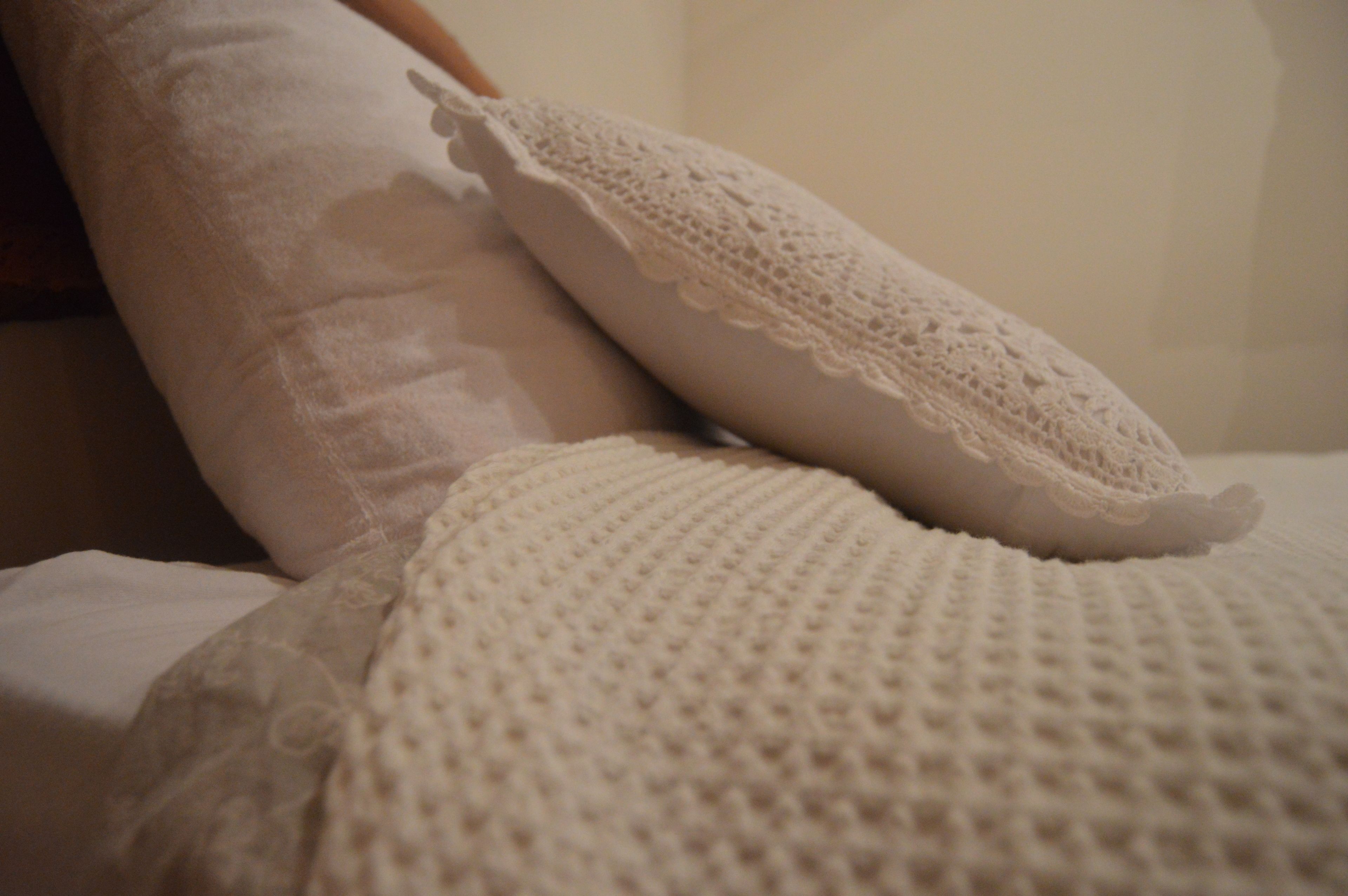 Una cama con una manta blanca de punto y un almohadón blanco con encaje.