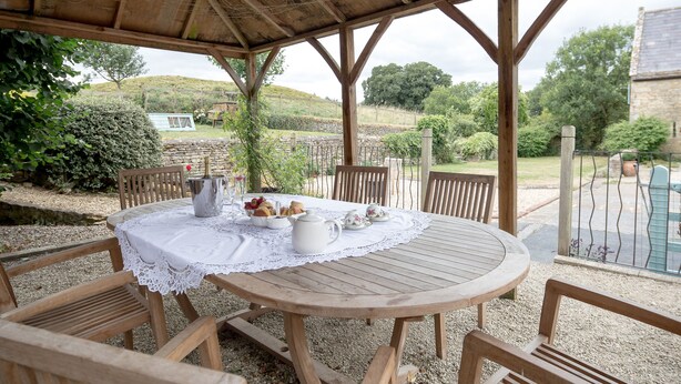 Une table en bois, recouverte d’une nappe en dentelle, entourée de chaises en bois, est placée sur une surface de gravier, sous une pergola en bois.