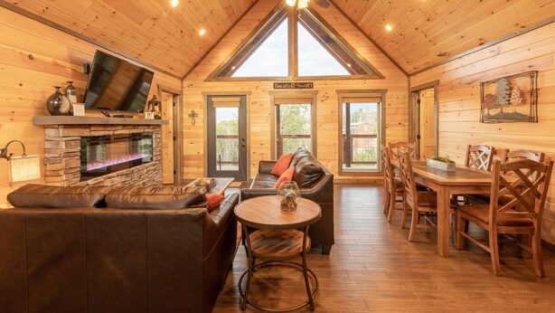 A cozy cabin interior with a stone fireplace, leather sofa, dining table, and a ceiling fan. -  Mt. LeConte Views! Cosy Luxury Cabin, Fire Pit, Hot Tub, Outdoor Pool, Location (Gatlinburg)