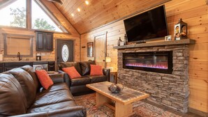 A cozy living room with a leather sofa, a stone fireplace, and a flat-screen TV. -  Mt. LeConte Views! Cosy Luxury Cabin, Fire Pit, Hot Tub, Outdoor Pool, Location (Gatlinburg)