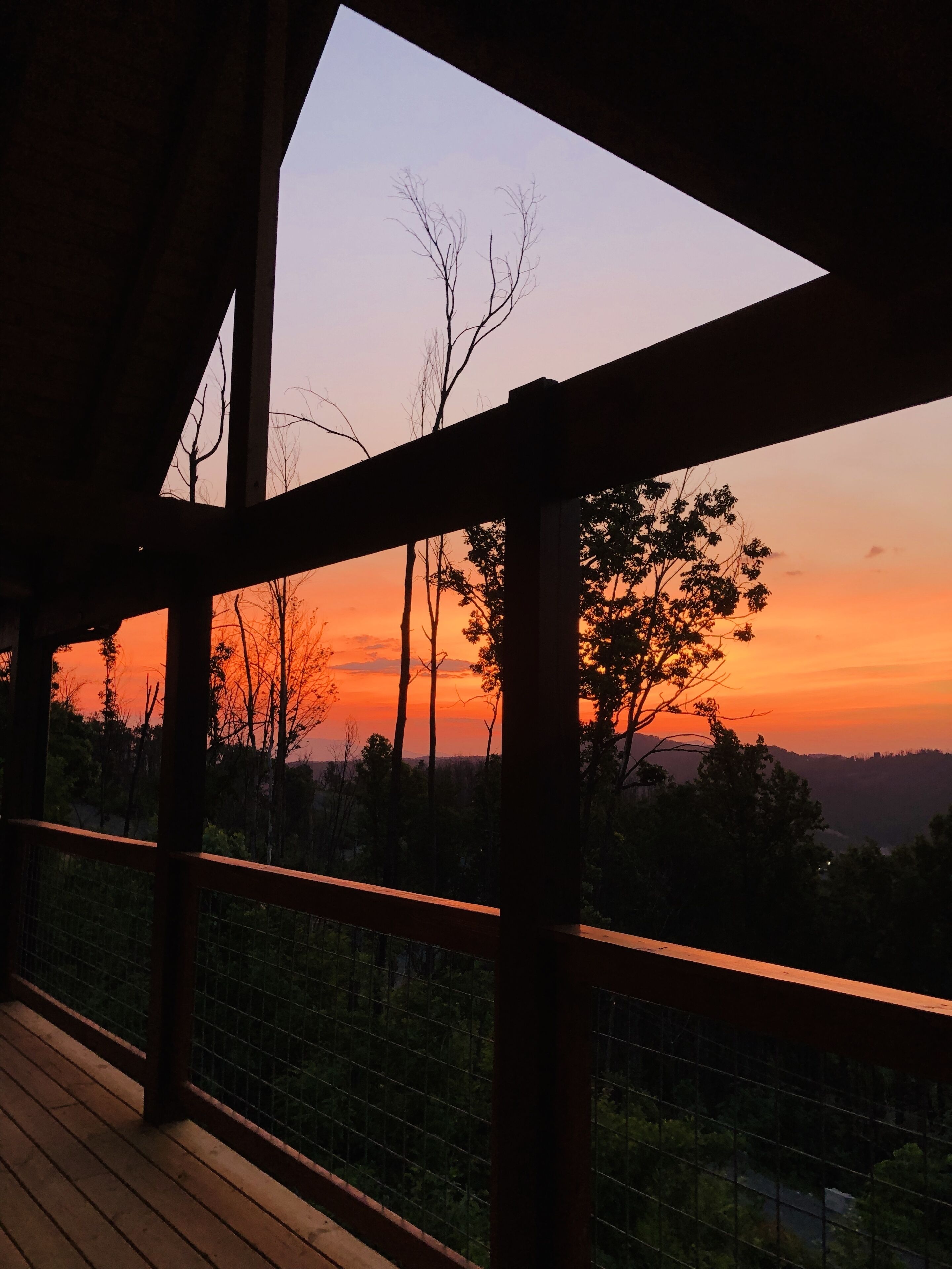 Blick von einer überdachten Terrasse mit Holzboden und Metallgeländer auf eine Landschaft mit Bäumen und Sonnenuntergang.