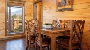 A wooden interior with a dining table and chairs, a window with a view of mountains, and a framed artwork on the wall. -  Mt. LeConte Views! Cosy Luxury Cabin, Fire Pit, Hot Tub, Outdoor Pool, Location (Gatlinburg)