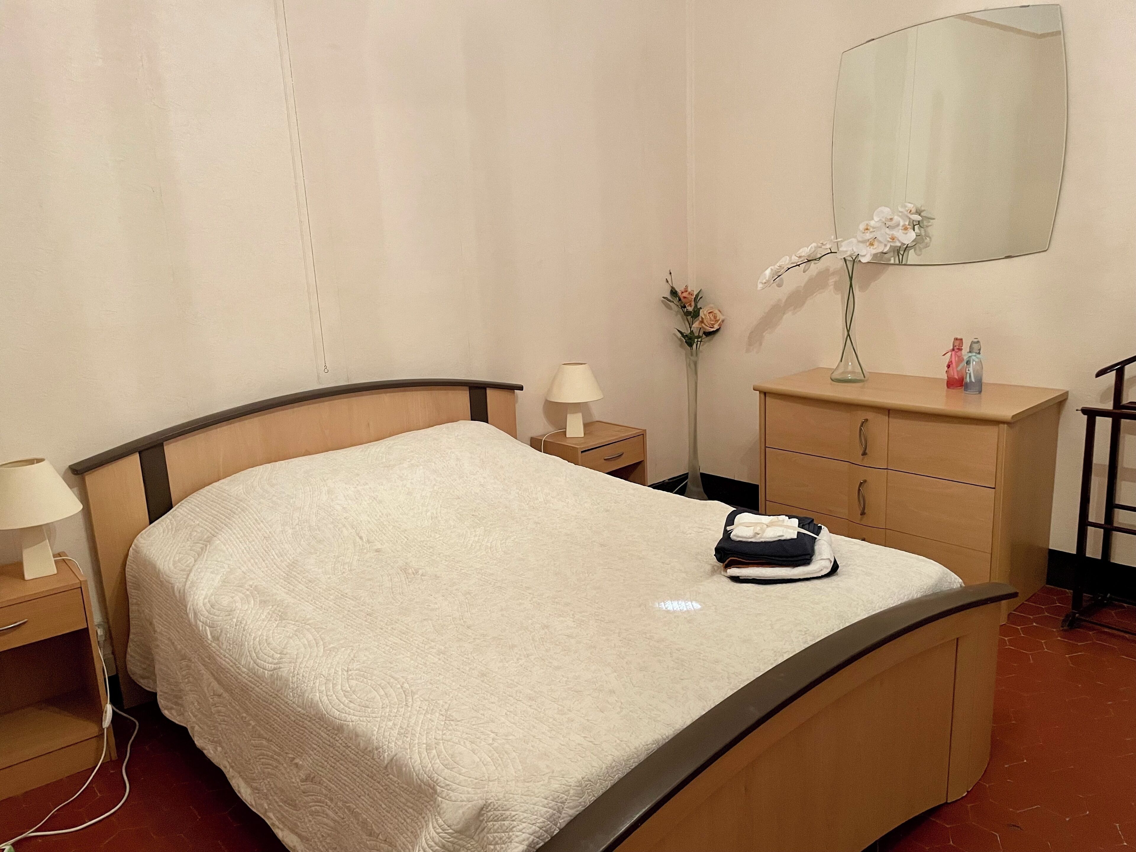A neatly made bed with a white quilt, a bedside table with a lamp, a dresser with a mirror, and a vase with flowers.