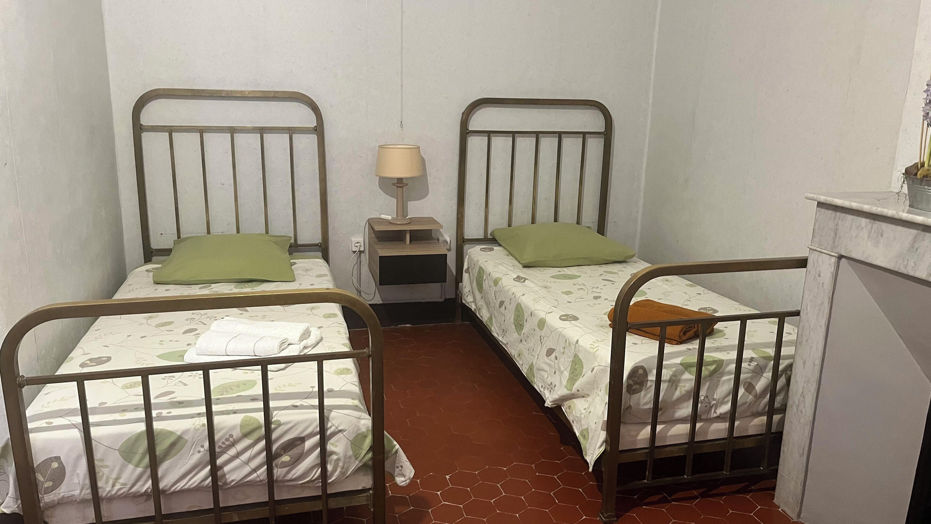 A small, dimly lit room with two metal bunk beds, each with a green pillow and a white bedspread. A small bedside table with a lamp and a power outlet is visible between the beds.